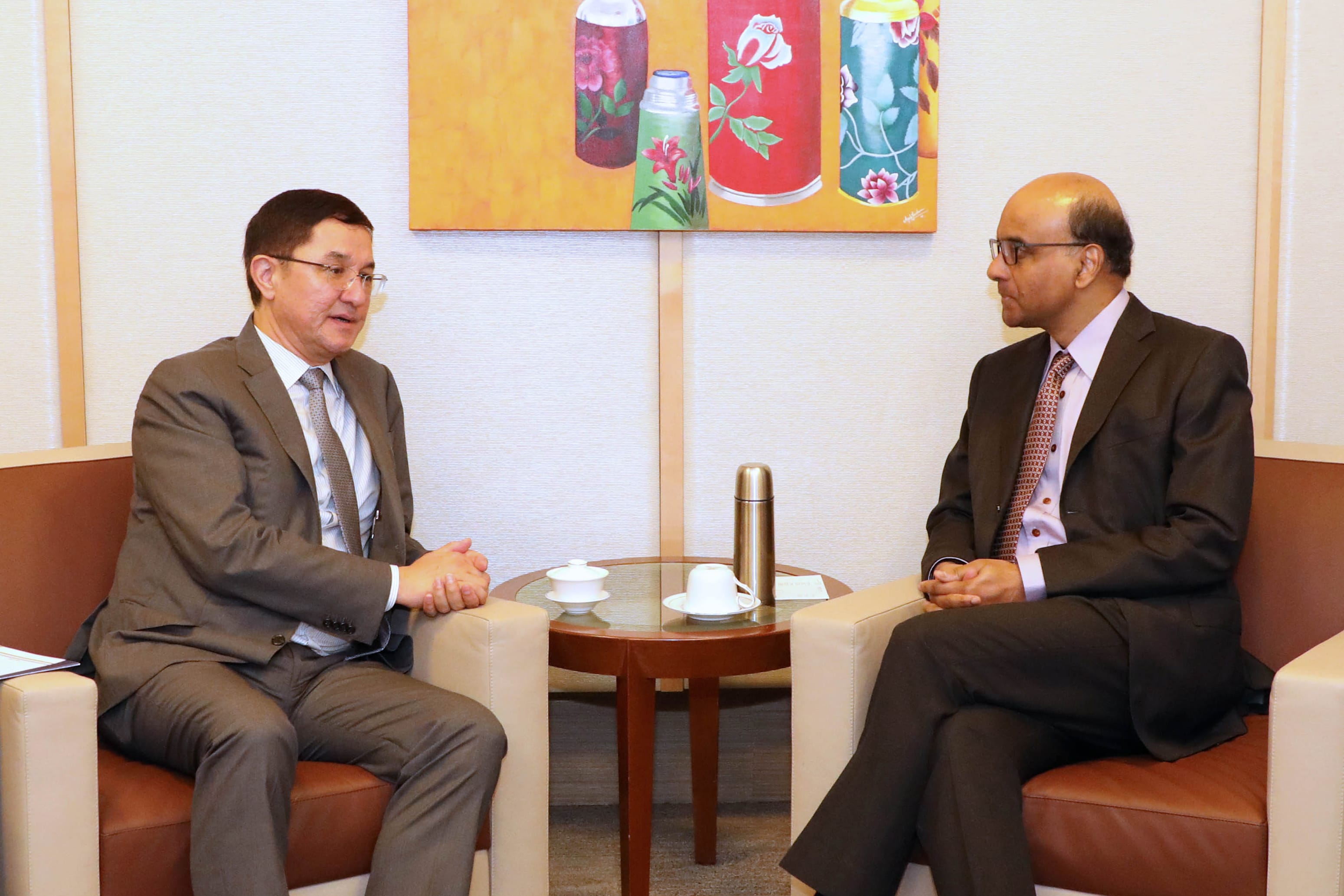 Two men in suits sit in armchairs facing each other with tea service on a table between them.